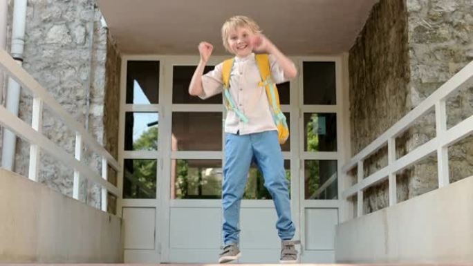 一个小男孩在学校大楼门口背着背包旋转跳跃。快乐的孩子完成课程后会很开心