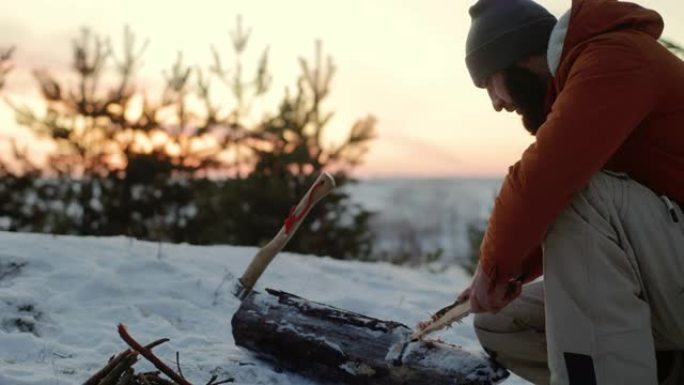 一名男子用刀准备木棉，以便在日落时在冬季森林中生火。冬季生存概念。