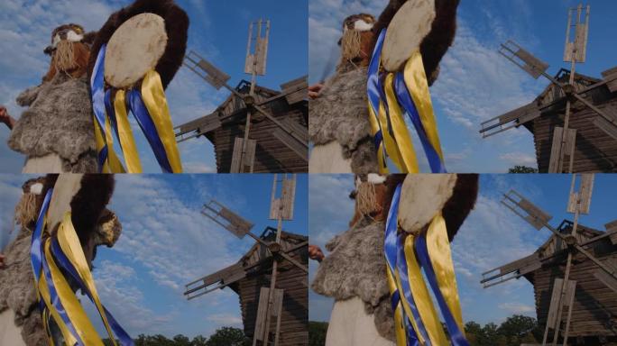 一个金发女孩穿着用兽皮制成的异教毛皮服装，戴着带角的山羊面具，在一个古老的木制风车和乡村风景的背景下