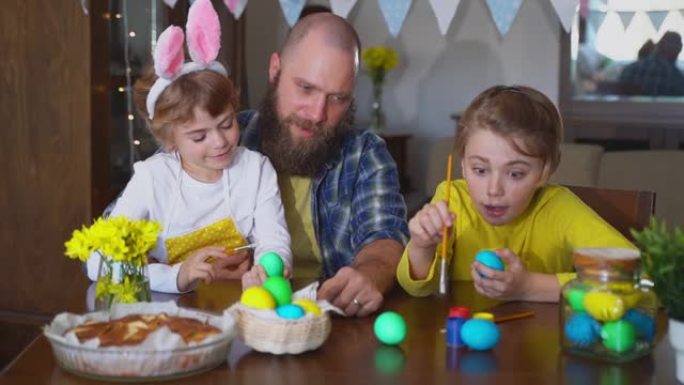 复活节家庭传统。父亲和两个白种人快乐的孩子，他们坐在家里的桌子上，用兔子耳朵染成鸡蛋，用油漆装饰鸡蛋