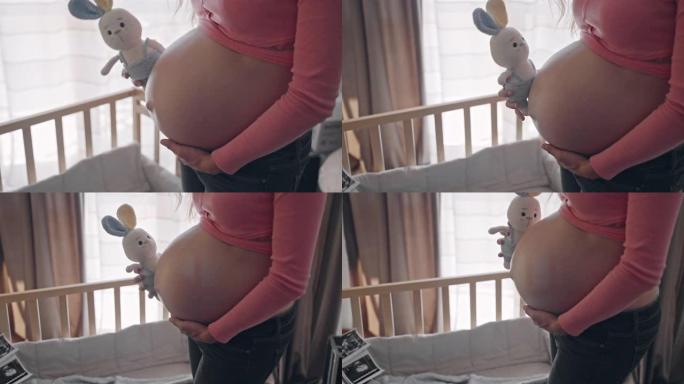 美丽的孕妇站在婴儿床旁边，手里拿着兔子玩具，快乐的母亲享受着产前护理她的肚子，孕妇期待着婴儿的孩子