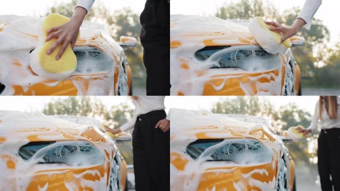 在户外自助洗车时，用黄色海绵洗车罩和汽车前灯的年轻白人妇女的手的特写裁剪图像。由泡沫覆盖的豪华车。户
