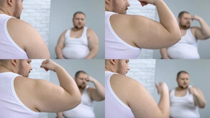沮丧的肥胖男性看着他虚弱的手臂肌肉，松弛的皮肤，脂肪团