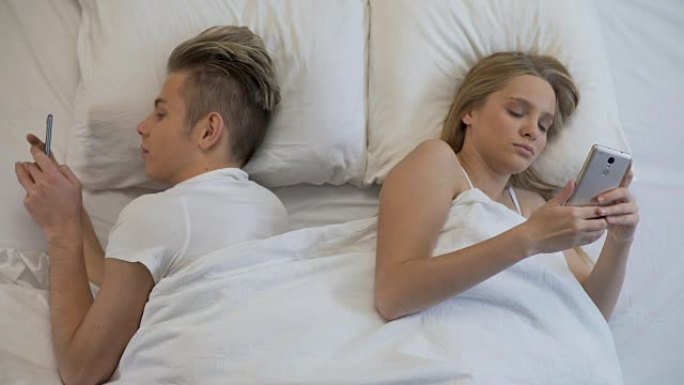 夫妇背靠背躺在床上，用智能手机，社交网络，忽略