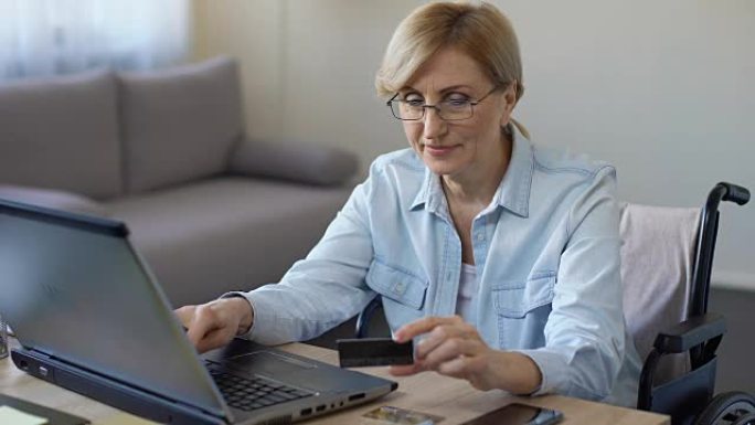 坐在轮椅上的快乐成年女性在笔记本电脑上输入卡号，在线购物