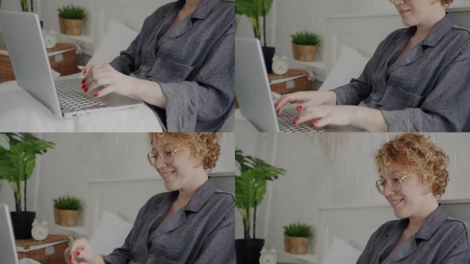 穿着睡衣的快乐女士的肖像在卧室的床上用笔记本电脑打字和大笑享受远程交流