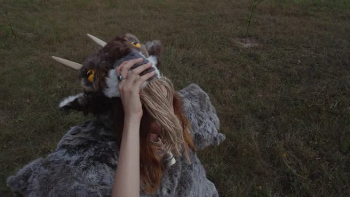 近距离拍摄金发女孩的肖像，她穿着一种异教动物般的皮毛，由皮肤制成，站在草地的背景下，戴着带角的山羊面