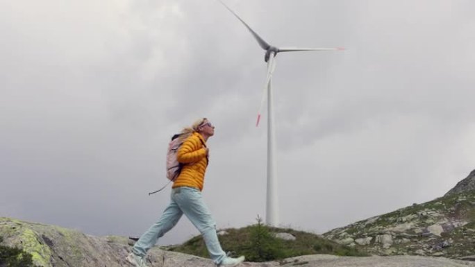 慢动作: 女人在山间小径上徒步旅行，背景式风力涡轮机发电站。可再生能源概念和对自然的热爱
