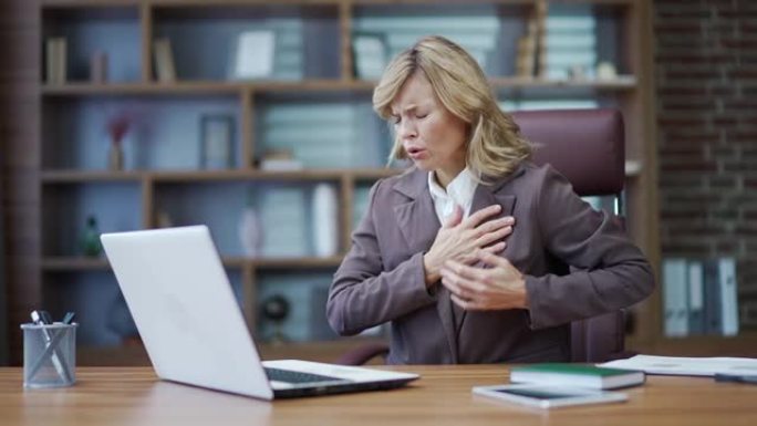 生病的成熟女商人在办公室的办公桌上工作时感到心痛。一个穿着西装的悲伤女人把手放在胸前