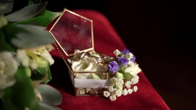玻璃盒子里有两个金色订婚戒指，红色表面有一个白色和蓝色花朵和叶子的树枝。订婚戒指，复制空间