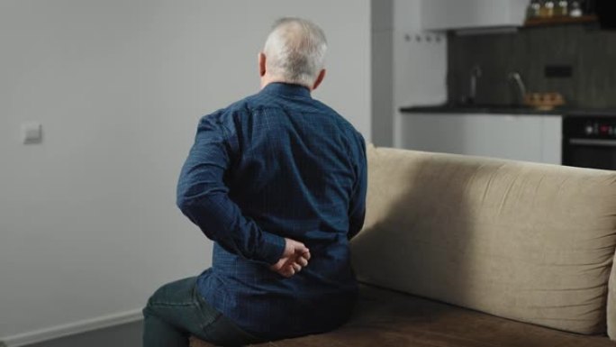 一位老人坐在家里的沙发上，背部疼痛。一个男人穿着衬衫在家，痛苦不堪