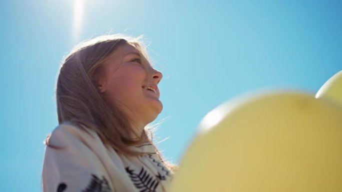 底角视图开朗的少女与黄色和蓝色的气球在户外阳光。在无云的夏日春天天空笑的背景下，快乐微笑的少年肖像。