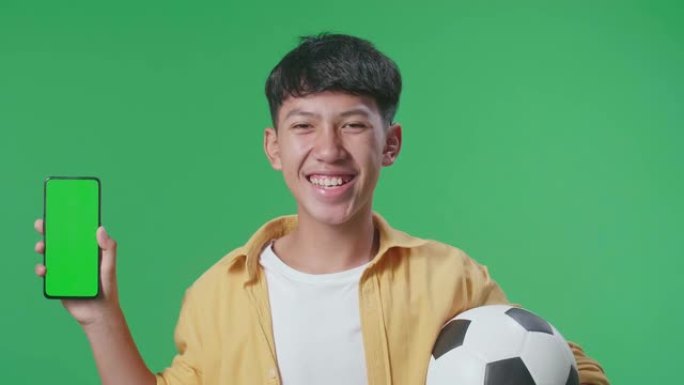 亚洲男孩的特写镜头显示模拟绿屏智能手机，并在绿屏背景上欢呼足球时摇摇头