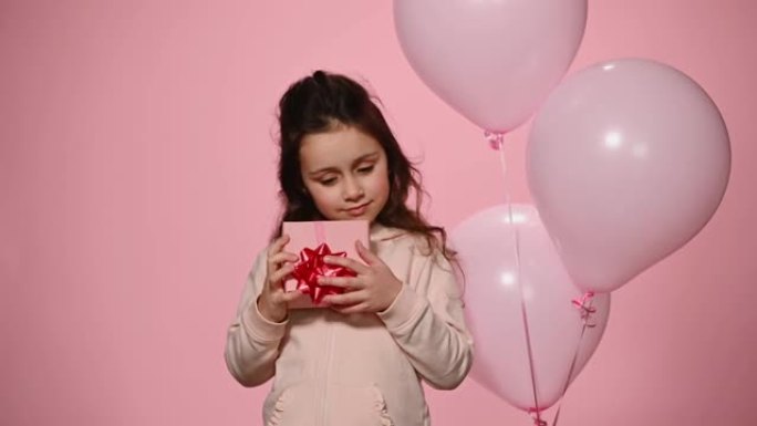 可爱的孩子摇晃礼品盒站在粉红色背景上的充气氦气球附近