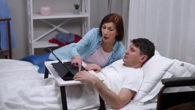 微笑的女人走进卧室，躺着生病的男人，用数字平板电脑在键盘上打字。不满意的高加索人激怒丈夫摇头，表情不