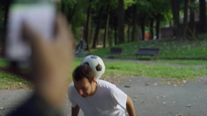 才华横溢的年轻足球运动员熟练地玩弄球，而他的朋友正在用智能手机拍摄他。时髦的人在公园里展示他的自由式