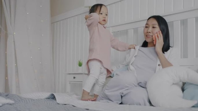 快乐的亚洲女士的慢动作在手机上说话，在家照顾床上的小孩