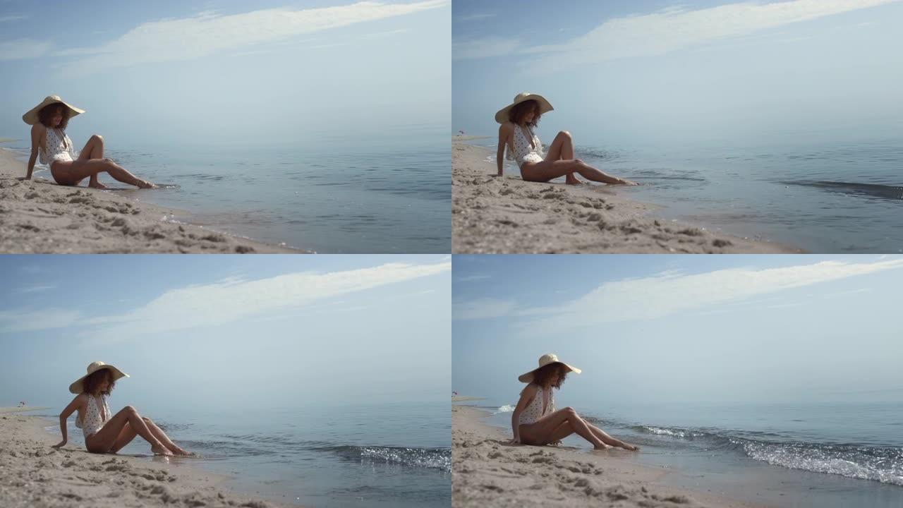 放松的女人坐在沙滩上洗沙在海浪中。女孩日光浴。