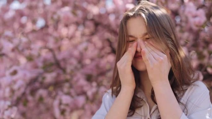 受刺激的高加索年轻女子站在盛开的樱花树附近，在户外的春天，抓挠发痒的眼睛。对花粉过敏反应。用于药品广