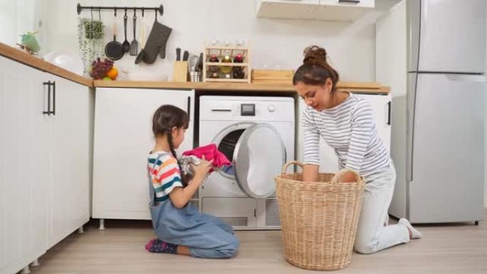 高加索美丽的母亲教小女儿洗脏衣服。可爱的小可爱女孩孩子帮助和学习父母妈妈把衣服放在家里的洗衣机里。家