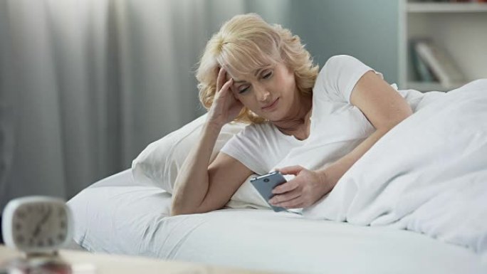 微笑的成熟女人躺在床上，早上在智能手机上查看电子邮件