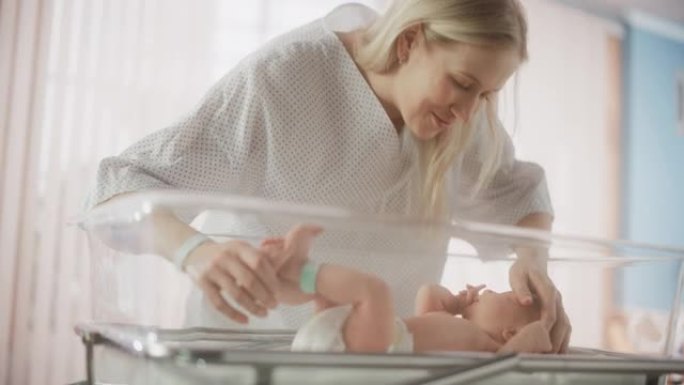 现代医院中母亲与新生婴儿联系的肖像。年轻女子在额头上亲吻她的孩子。婴儿躺在产科诊所的摇篮中
