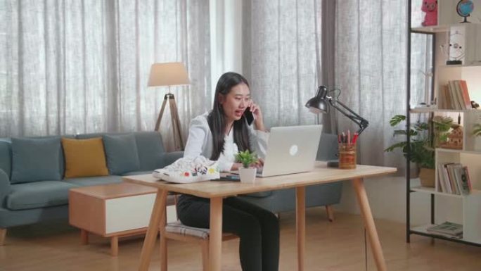 亚洲女鞋设计师在家中使用笔记本电脑时在智能手机上聊天