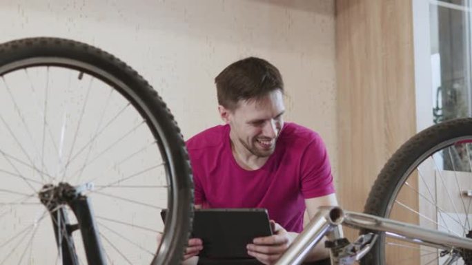 带平板电脑的人坐在自行车附近，踏板被拆下。快乐的家伙看着自行车。