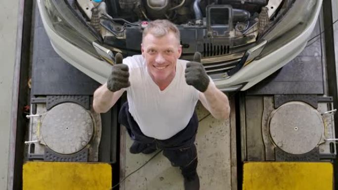 顶视图。专业汽车机械师或技术人员的肖像在车库检查和修理内部部件发动机系统。微笑向镜头竖起大拇指。汽车
