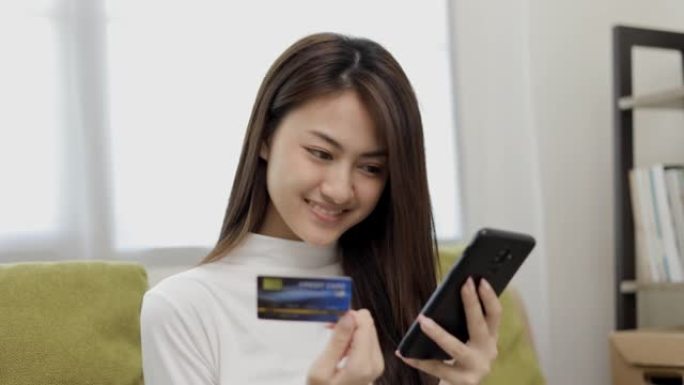 网上购物。年轻的亚洲少女将信用卡的序列号输入智能手机进行购物付款。在家在线送货购买商品。微笑的女性感