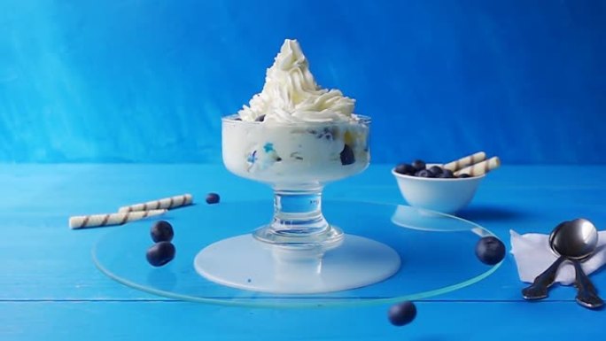 蓝莓落入玻璃冰淇淋杯中，该杯在浅蓝色背景下的蓝色木制桌子上的玻璃托盘上旋转。具有加速、减速效果的慢动