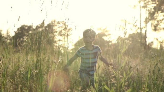 慢动作小男孩在夏日的草地上奔跑，带着美丽的日落光线穿过高高的草地。童梦，快乐的童年，自由的观念。
