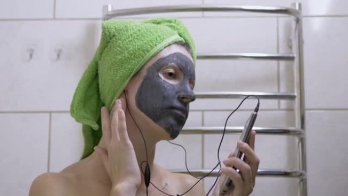 脸上戴着粘土面具的女孩在智能手机上戴着耳机听音乐