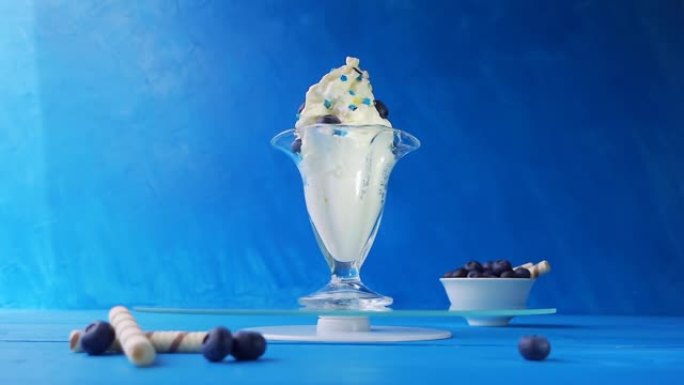 在一个美丽的玻璃玻璃杯中加入蓝莓的冰淇淋在淡蓝色背景上的蓝色木制桌子上的玻璃托盘上旋转。具有加速，减
