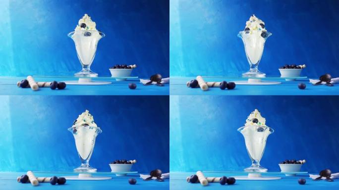 在一个美丽的玻璃玻璃杯中加入蓝莓的冰淇淋在淡蓝色背景上的蓝色木制桌子上的玻璃托盘上旋转。具有加速，减