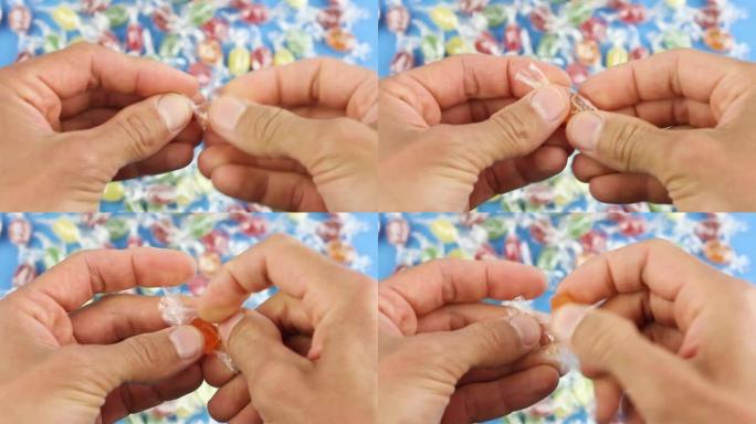 双手从透明包装纸上展开一个橙色糖果，在许多彩色糖果，糖糖果的背景下