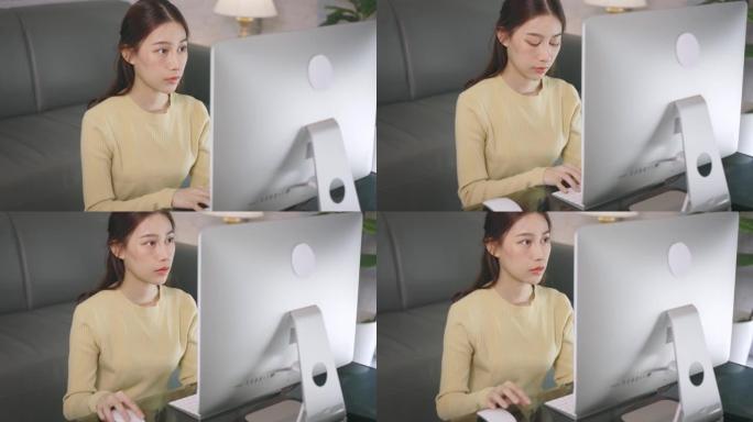 年轻的亚洲女商人在家工作，坐在客厅里用台式电脑在键盘上打字