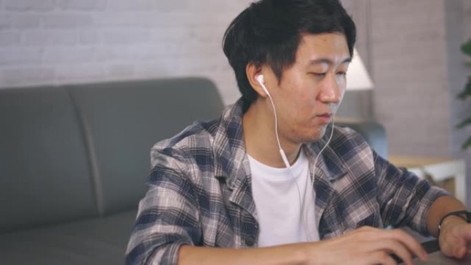 穿着休闲服装和耳机的年轻亚洲男性企业家深夜在家在电脑笔记本电脑上工作