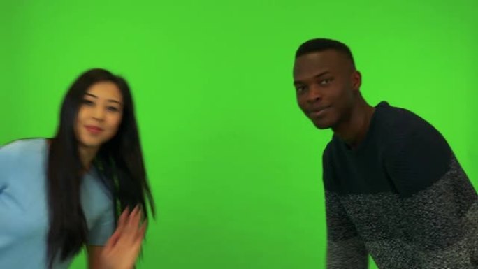 一名年轻的亚洲女子和一名年轻的黑人男子靠在镜头前，微笑着对着镜头挥手 -- 绿屏工作室