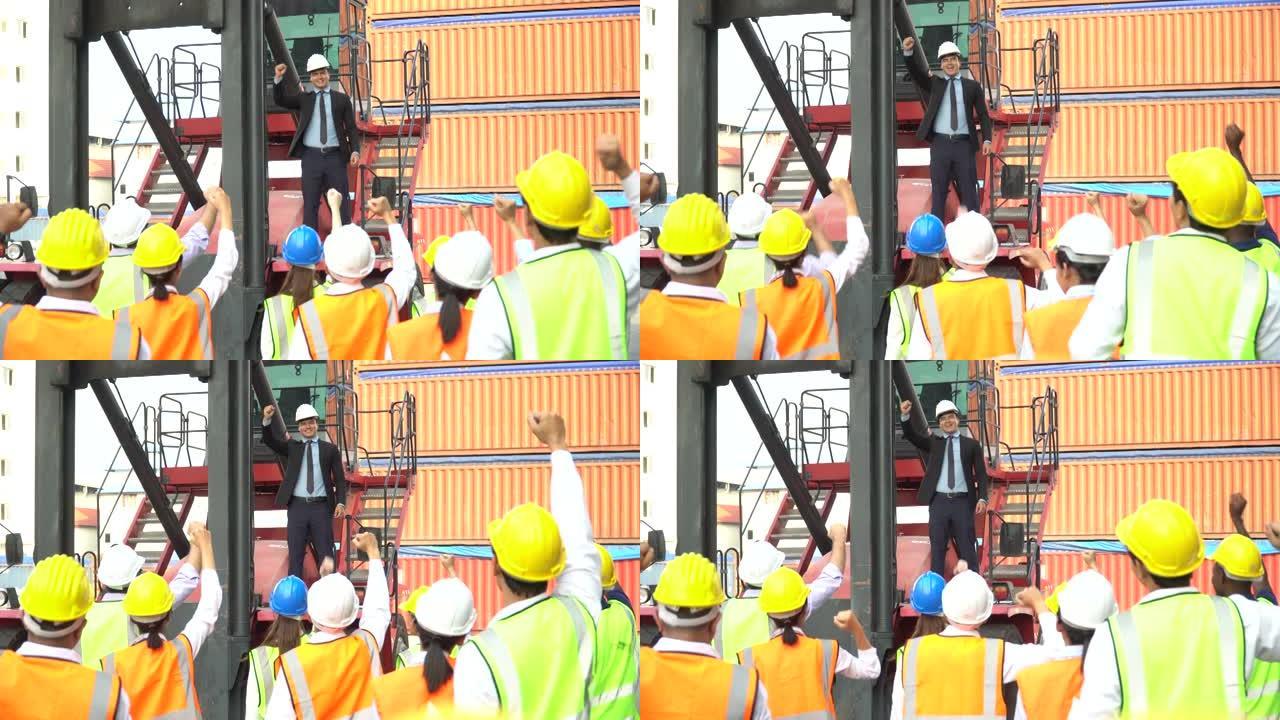 上午的会议上，一名商人站在叉车上向集装箱工人演讲，激动地举起手来祝贺成功。一群劳动庆祝在货轮港口的仓