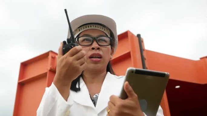 戴着白色头盔的女工程师拿着数字平板电脑，在大坝施工现场使用无线电通信发电。清洁能源和技术概念。