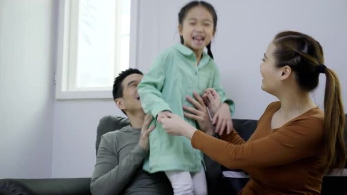 亚洲家庭父母和女儿看电视回家开心笑。女孩和父母一起享受在家玩耍的空闲时间。周末活动幸福家庭生活理念。