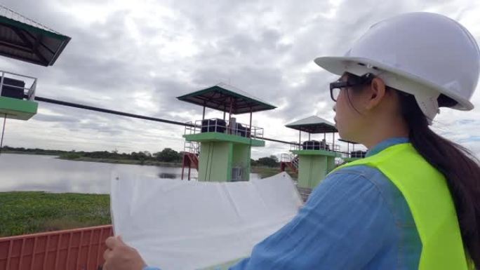 穿着白色头盔和黄色制服的女工程师在蓝图中读取数据，并检查大坝施工现场的水系统以发电。