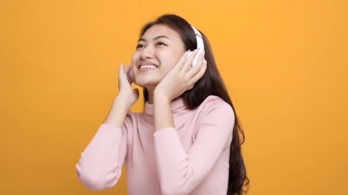 开朗的亚洲女少年用白色耳机在孤立的黄色背景上跳舞听音乐。美丽的年轻女子手触摸无线耳机，享受音乐的乐趣