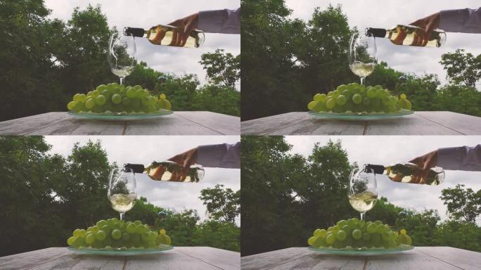 在大自然的背景下，将白葡萄酒从瓶子里倒入玻璃杯中。玻璃托盘上的一杯白葡萄酒，木桌上放着一根绿色葡萄的