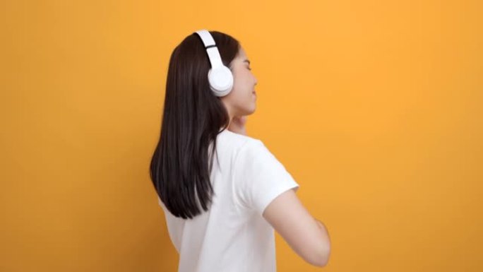 开朗的亚洲女少年用白色耳机在孤立的黄色背景上跳舞听音乐。美丽的年轻女子手触摸无线耳机，享受音乐的乐趣