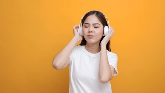 开朗的亚洲女少年用白色耳机在孤立的黄色背景上跳舞听音乐。美丽的年轻女子手触摸无线耳机，享受音乐的乐趣