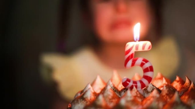 女孩看着蛋糕上有数字5的燃烧蜡烛，许愿。生日，五周年，五年，蜡烛火焰，橙色火焰。儿童假期。特写