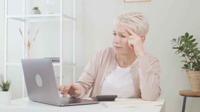沮丧的高级女性坐在桌子旁，拿着笔记本电脑，应对财务压力，因退休付款延迟而感到压力，担心很多或对金钱感