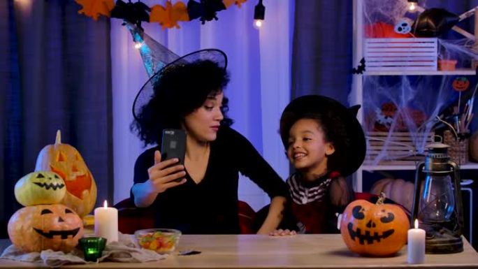 小女孩和年轻女子戴着巫婆帽子的非洲裔美国人使用智能手机通过视频通话进行交流。妈妈和女儿坐在万圣节之夜